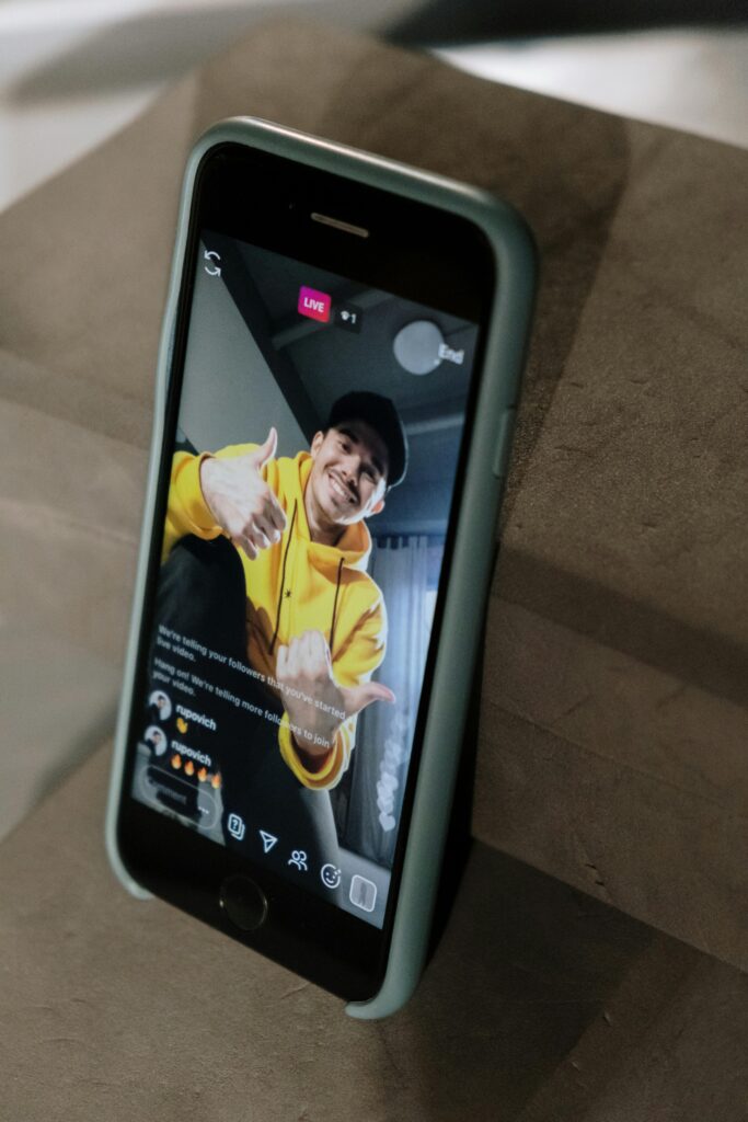 A man in a yellow hoodie engaging in a live stream on a smartphone display, indoors.